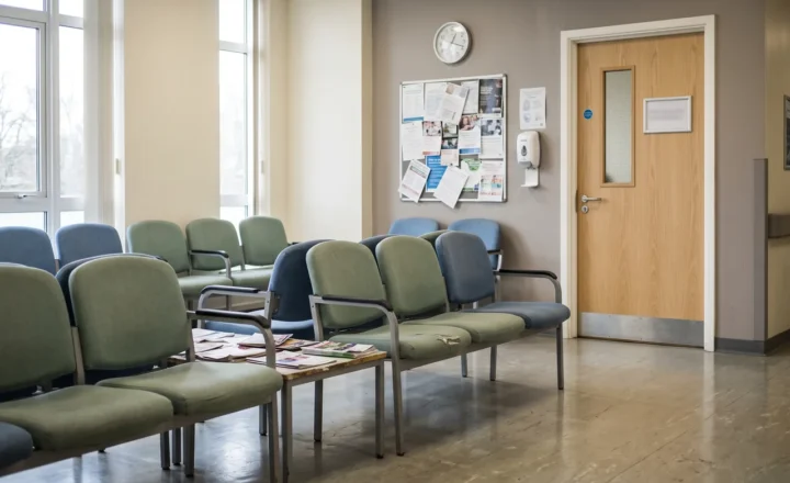 An empty waiting room with rows of green and blue chairs, a low table covered with magazines, and tall windows on the left. A clock, noticeboard and wall dispenser hang beside a closed wooden door.