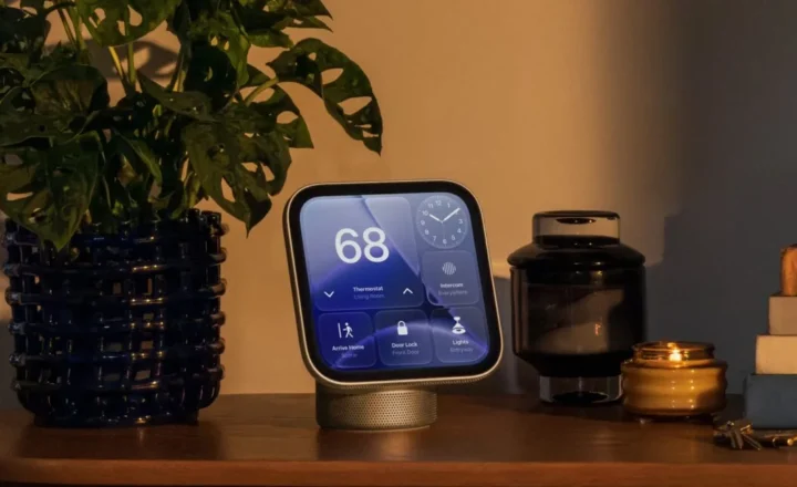 A rounded-square smart display on a cylindrical speaker base sits on a wooden tabletop. Its screen shows a thermostat reading of 68, an analog clock and smart-home controls. A leafy plant, dark glass vessel, lit candle, stacked books and keys surround it.