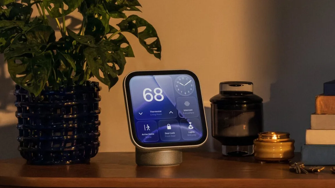 A rounded-square smart display on a cylindrical speaker base sits on a wooden tabletop. Its screen shows a thermostat reading of 68, an analog clock and smart-home controls. A leafy plant, dark glass vessel, lit candle, stacked books and keys surround it.