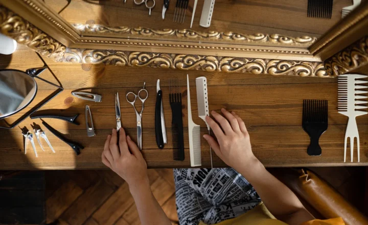 Overhead view of hands arranging scissors, combs, hair clips, and a straight razor on a wooden table beneath an ornate gold-colored mirror.