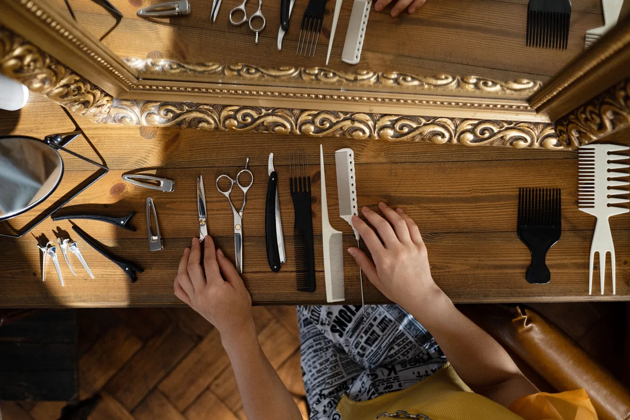 Overhead view of hands arranging scissors, combs, hair clips, and a straight razor on a wooden table beneath an ornate gold-colored mirror.
