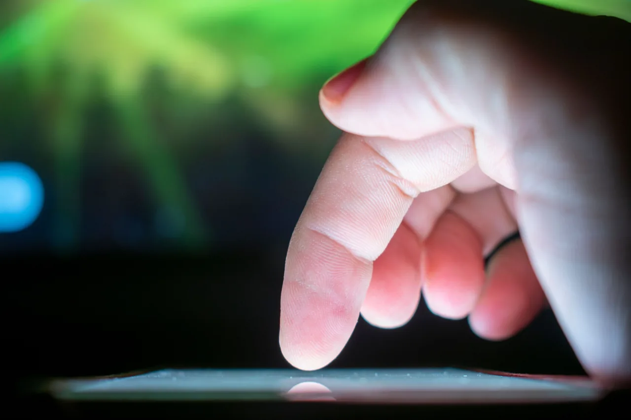 Close-up of a hand with a fingertip poised above an illuminated touchscreen against a blurred background.
