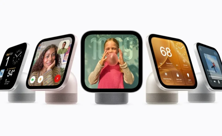 Five smart displays with rounded black screens and perforated speaker bases on a white background. The screens show a clock and weather, a video call, a child blowing a bubble, smart home controls and music playback.
