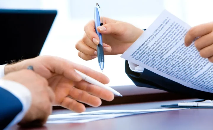 Close-up of hands holding pens and reviewing a printed document at a desk, with a laptop and clipboard nearby.
