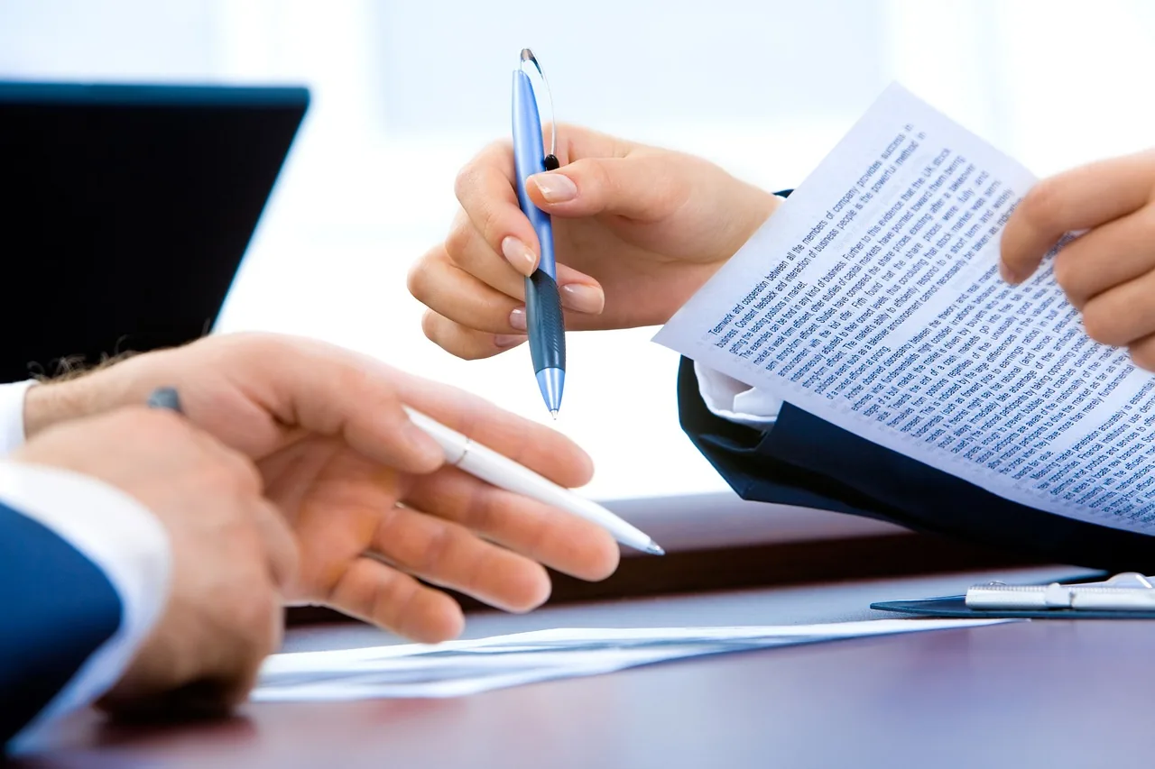 Close-up of hands holding pens and reviewing a printed document at a desk, with a laptop and clipboard nearby.