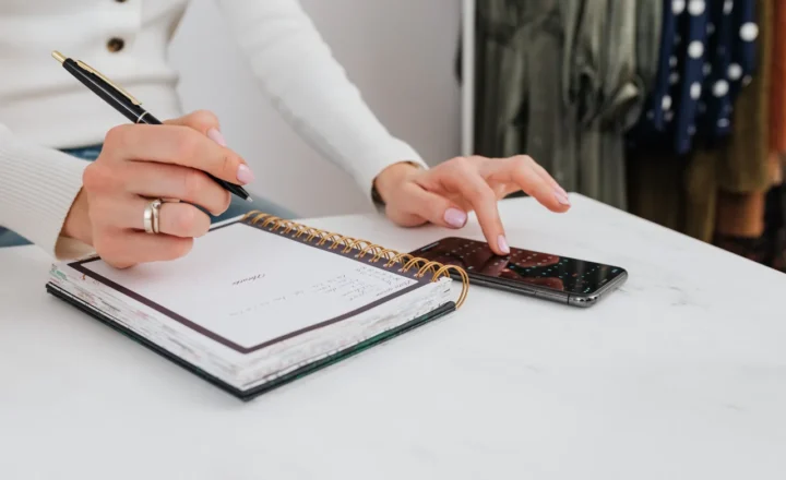 Close-up of hands holding a pen over a spiral notebook and tapping a smartphone on a white table.