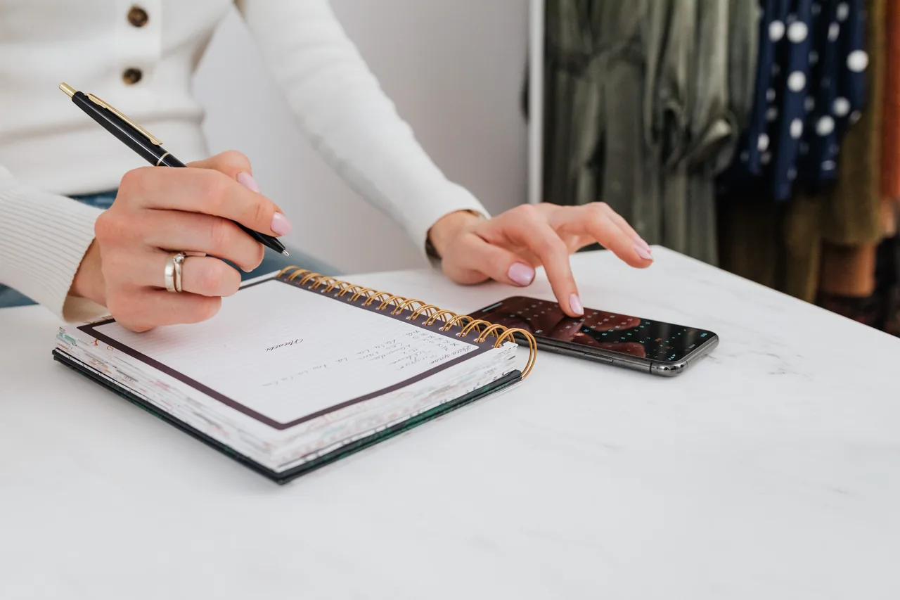 Close-up of hands holding a pen over a spiral notebook and tapping a smartphone on a white table.