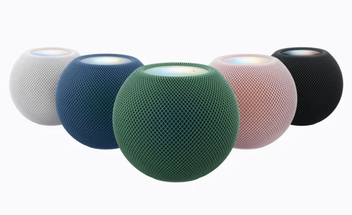 Five spherical mesh-covered smart speakers in white, blue, green, pink, and black on a white background, with illuminated circular touch panels on top. The green speaker sits prominently in front.