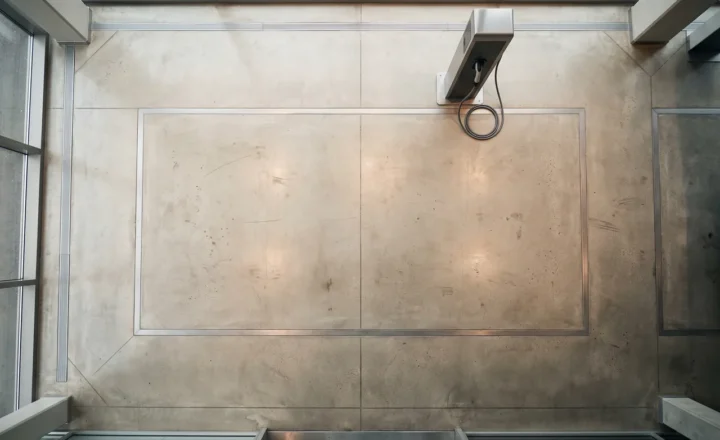 Overhead view of an empty concrete parking bay outlined by metal strips, with a charging unit and looped black cable near the upper edge. Glass panels and structural beams frame the space.