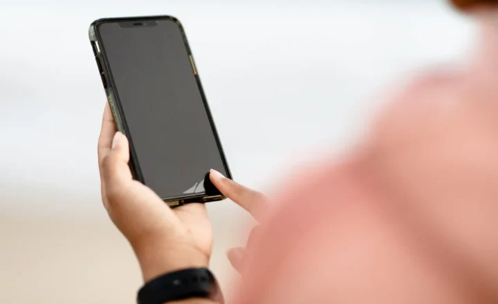 Close-up of hands holding a smartphone with a dark screen, with one finger touching its lower edge and a blurred pink-clad shoulder in the foreground.
