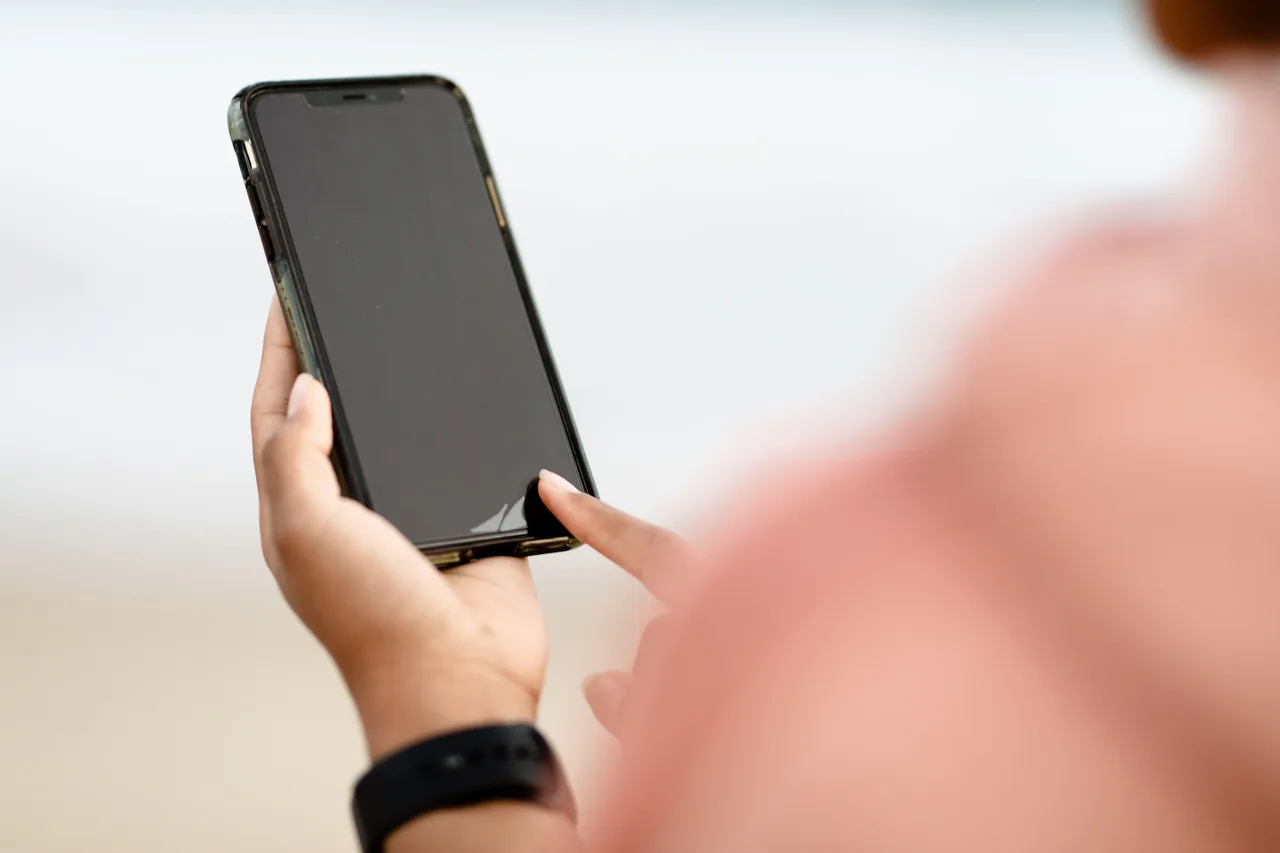 Close-up of hands holding a smartphone with a dark screen, with one finger touching its lower edge and a blurred pink-clad shoulder in the foreground.