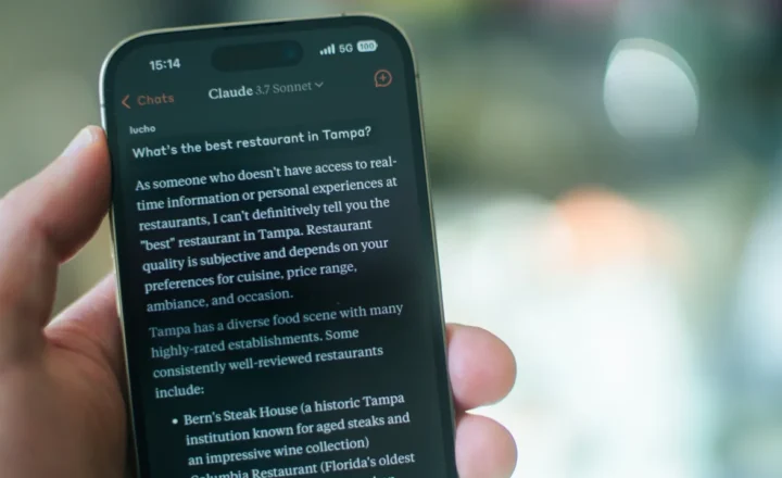 A hand holds a smartphone displaying a Claude 3.7 Sonnet conversation about restaurant recommendations in Tampa against a blurred background.