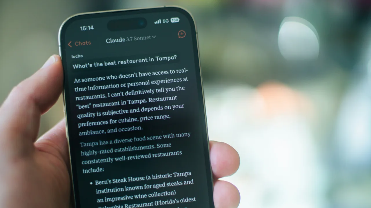 A hand holds a smartphone displaying a Claude 3.7 Sonnet conversation about restaurant recommendations in Tampa against a blurred background.