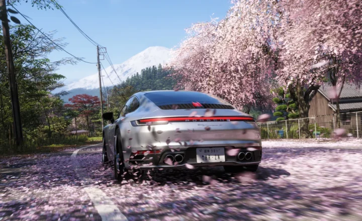 Rear view of a silver Porsche driving along a road covered in pink petals, with flowering cherry trees, wooden buildings, utility lines, and a snow-covered mountain in the background.