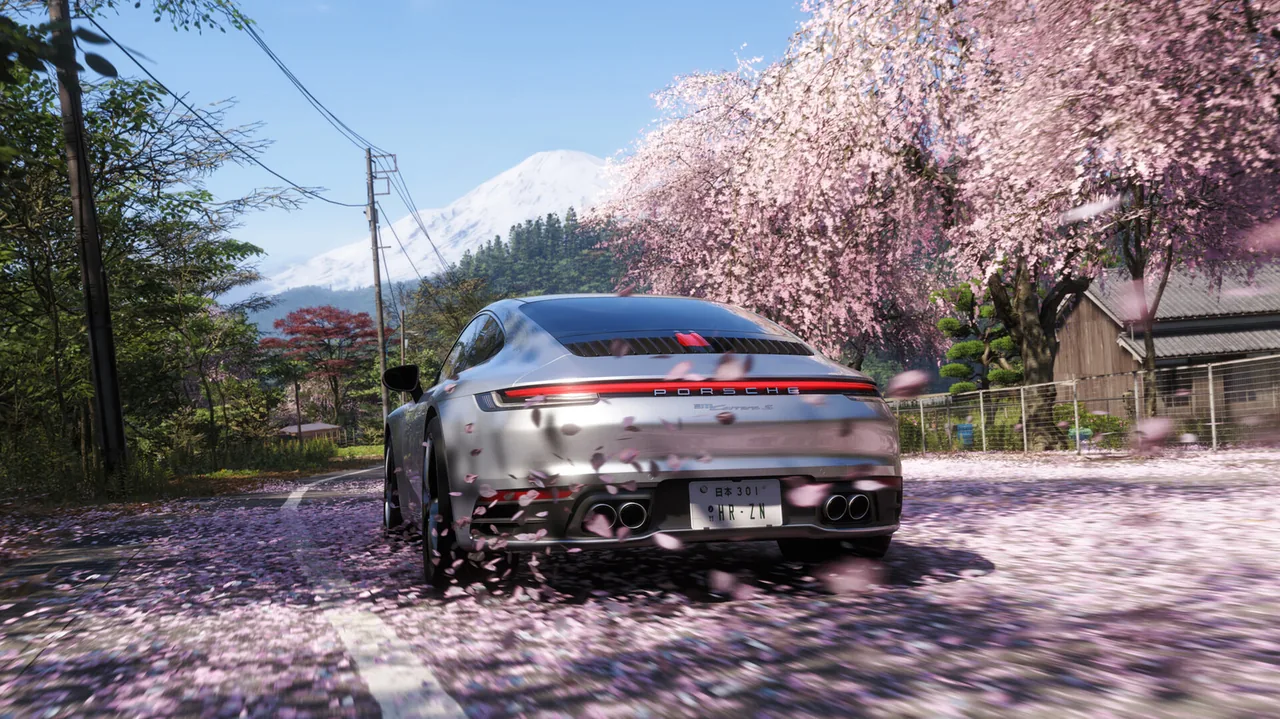 Rear view of a silver Porsche driving along a road covered in pink petals, with flowering cherry trees, wooden buildings, utility lines, and a snow-covered mountain in the background.